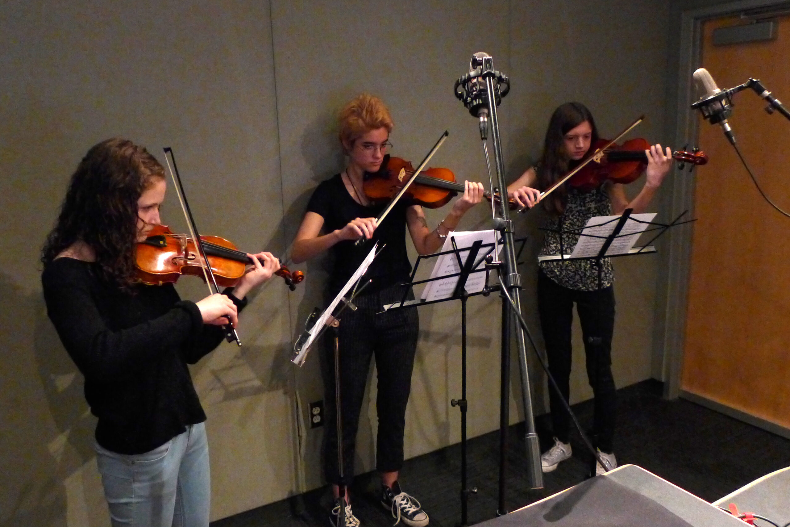 Three teens playing violin looking at sheet music on music stands in a radio studio