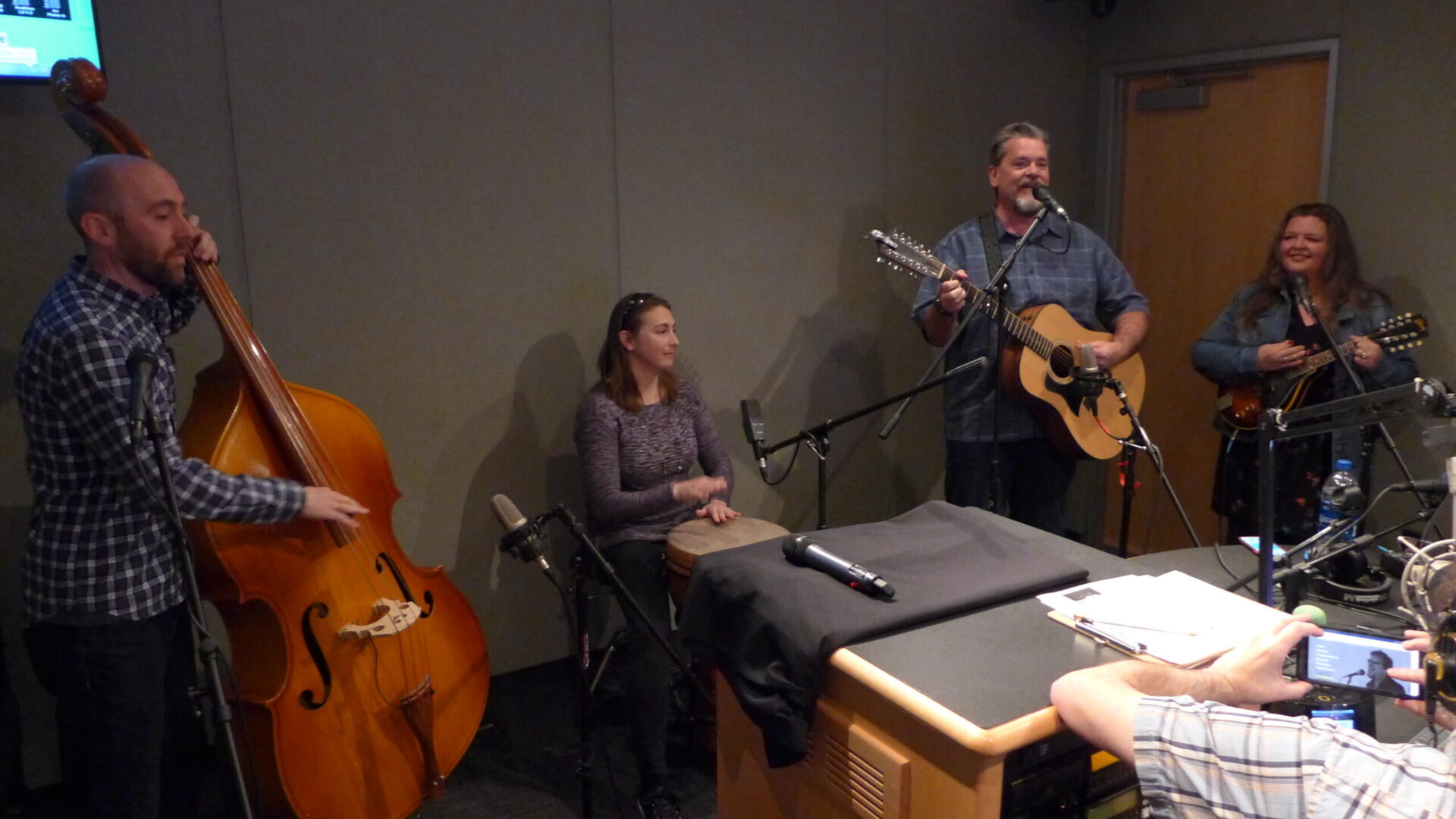 An adult playing upright bass, an adult playing a drum, an adult playing a guitar and singing into a microphone, and an adult playing a mandolin in a radio studio