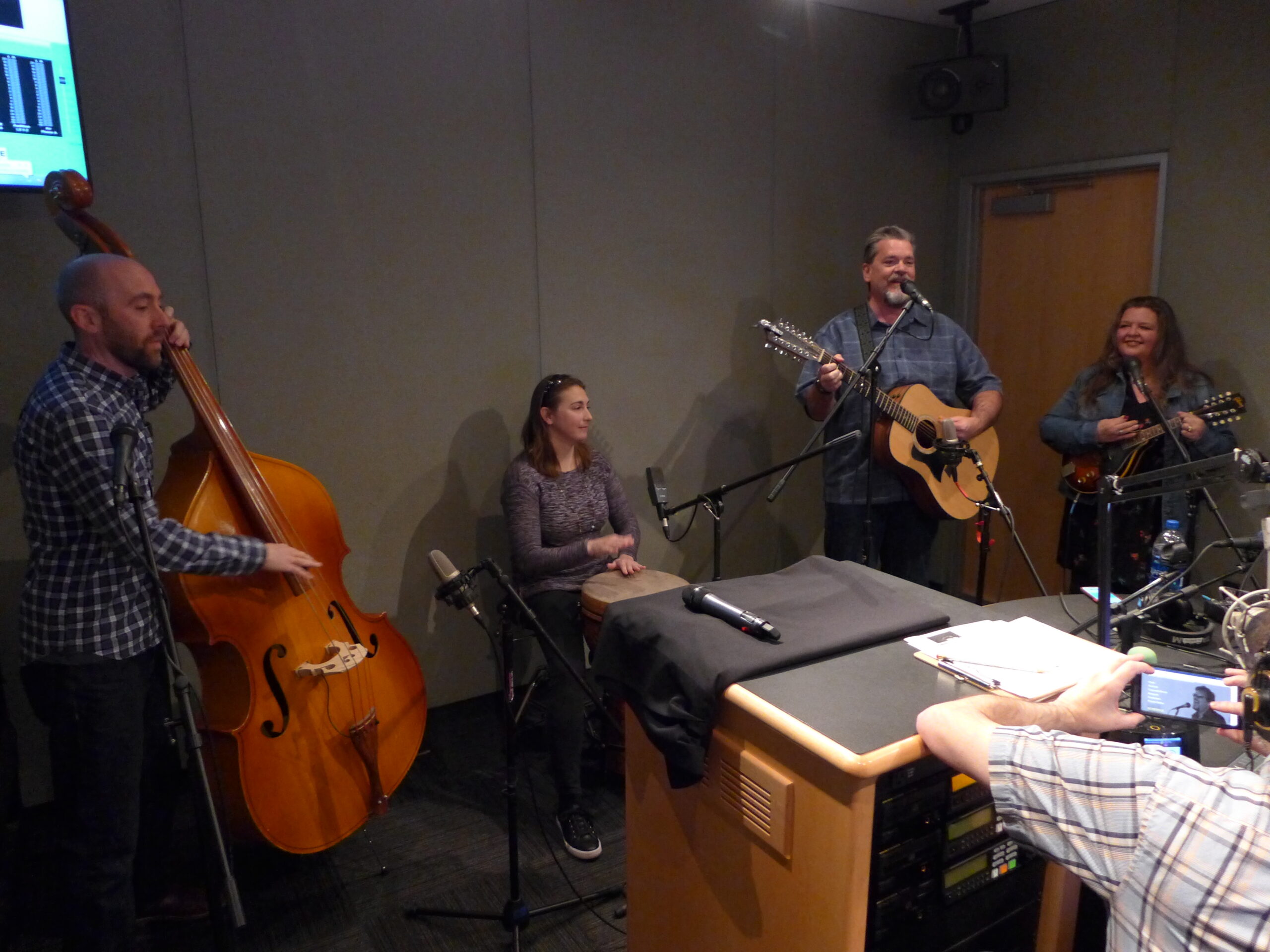 An adult playing upright bass, an adult playing a drum, an adult playing a guitar and singing into a microphone, and an adult playing a mandolin in a radio studio
