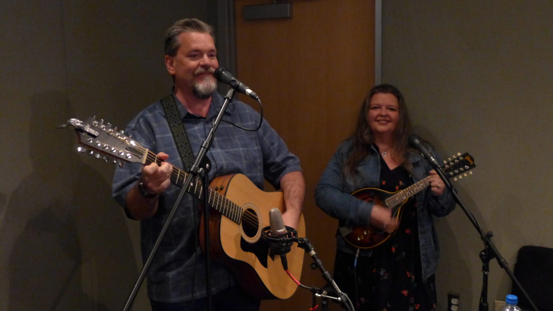 An adult holding a guitar in front of microphones and an adult holding a mandolin in front of microphones in a radio studio