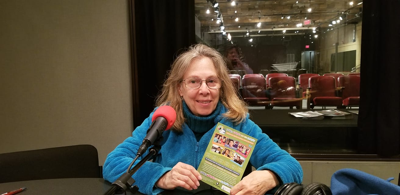 Adult wearing glasses sitting behind a microphone holding a pamphlet in a radio studio