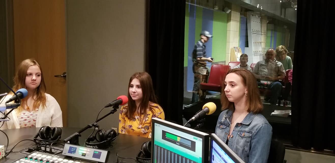 Three teens sitting behind microphones in a radio studio