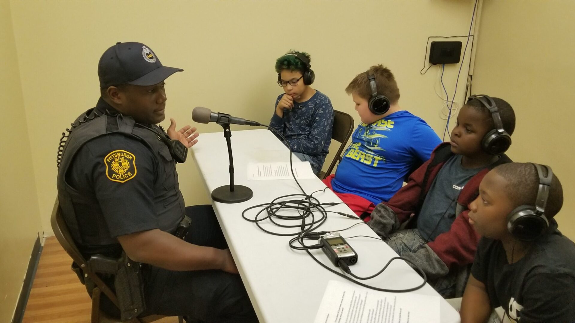 Five youth wearing headphones interviewing an adult wearing a police uniform speaking into a microphone connected to a portable recorder