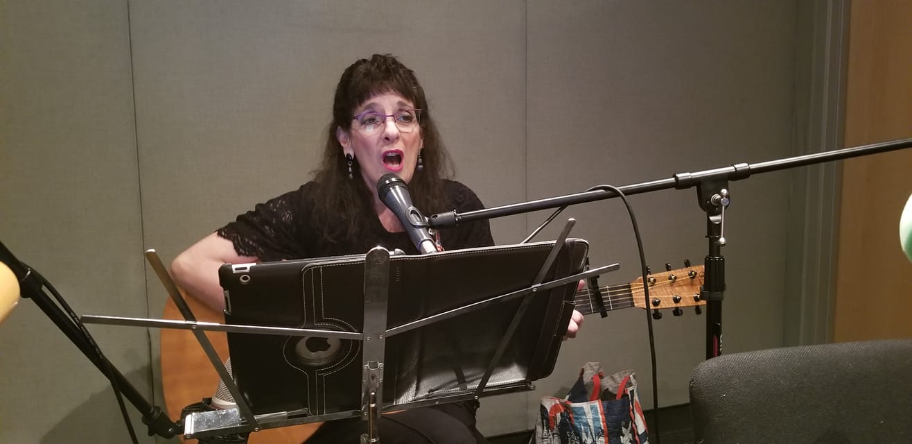 Adult playing guitar singing into a microphone and looking at a tablet on a music stand in a radio studio