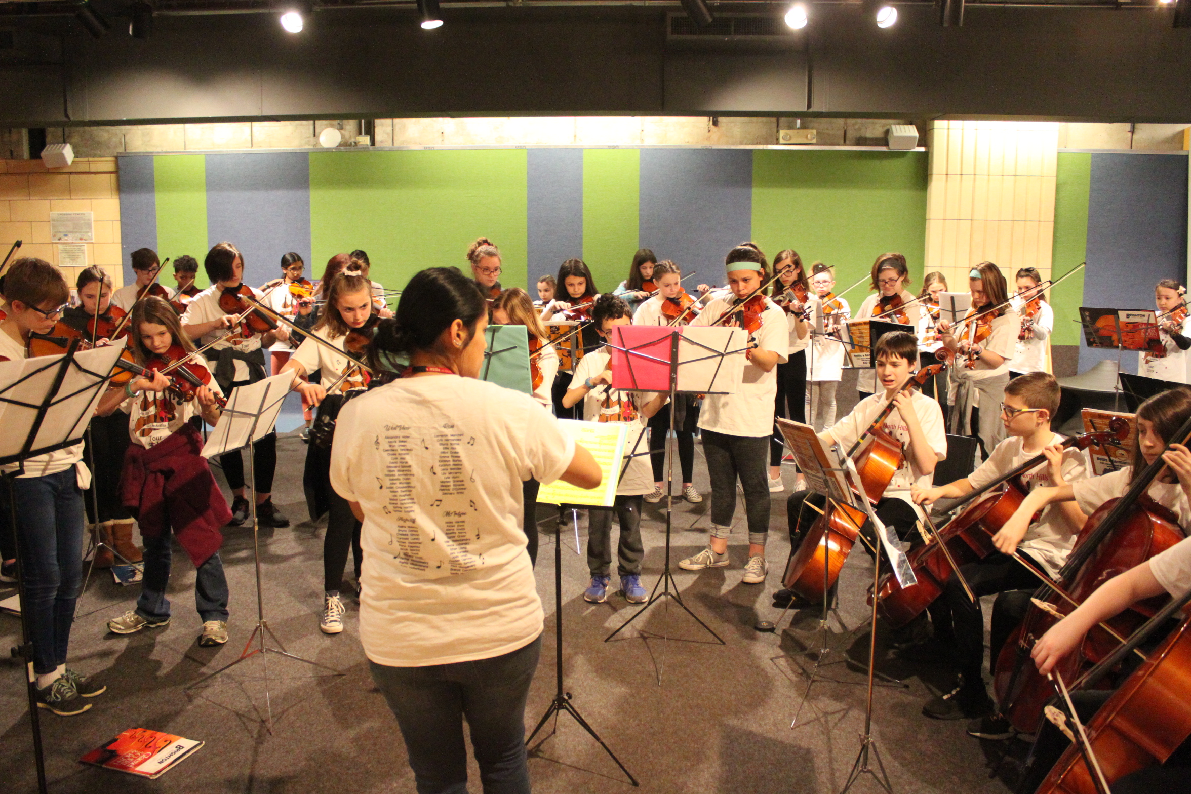 An adult looking at sheet music on a music stand conducting a large group of youth in an orchestra