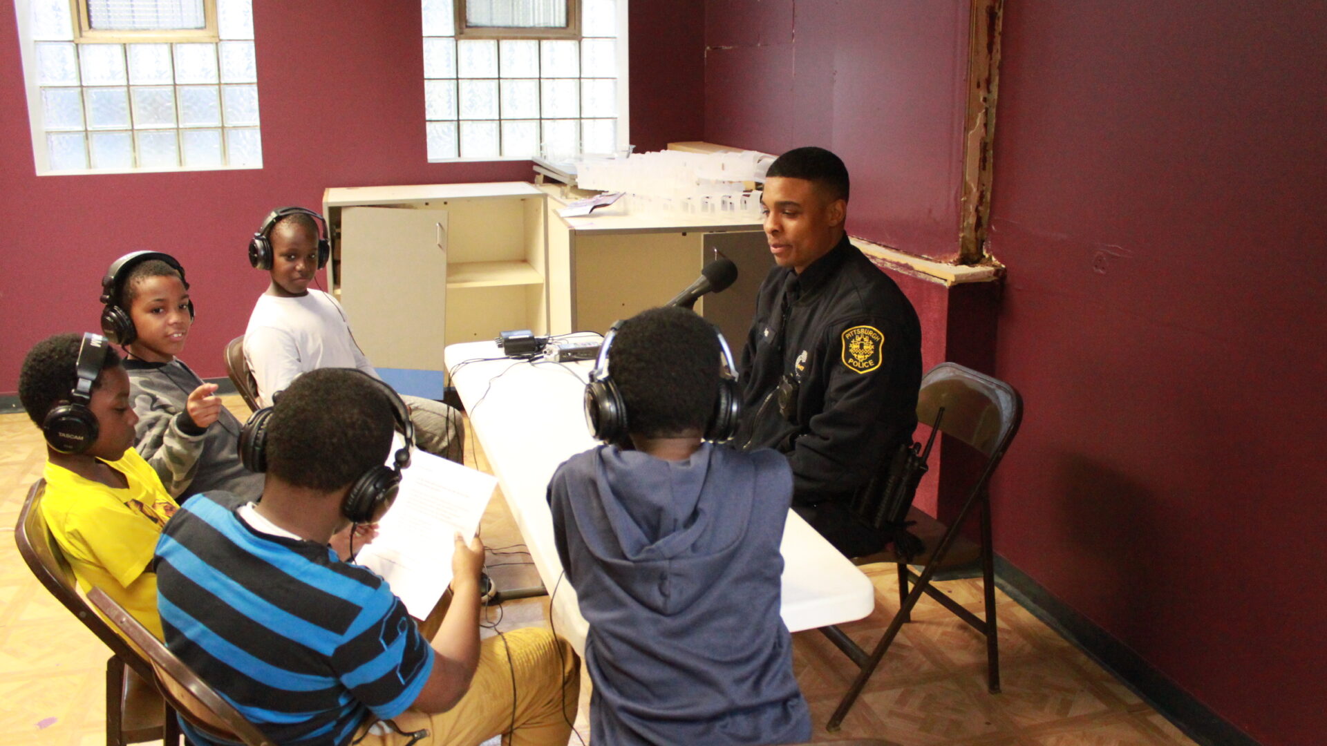 Five children wearing headphones interviewing an adult wearing a police uniform speaking into a microphone connected to a portable recorder