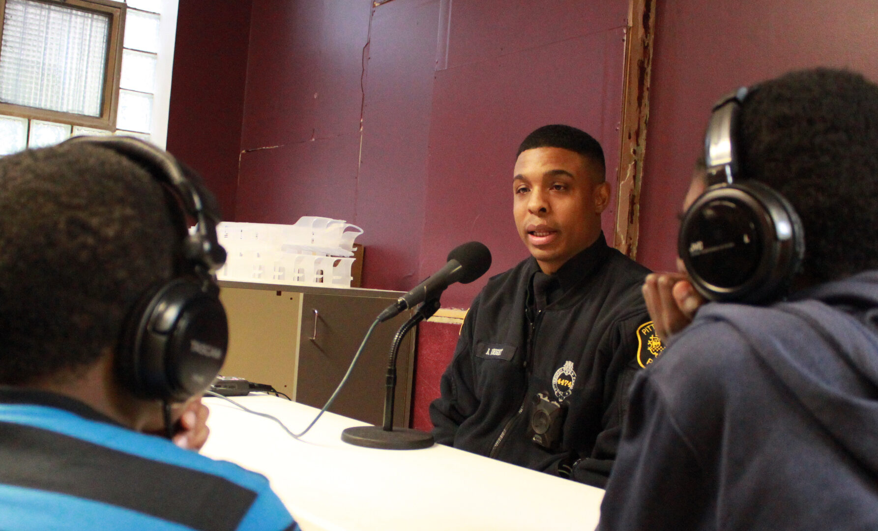 Two youth wearing headphones interviewing an adult wearing a police uniform speaking into a microphone connected to a portable recorder