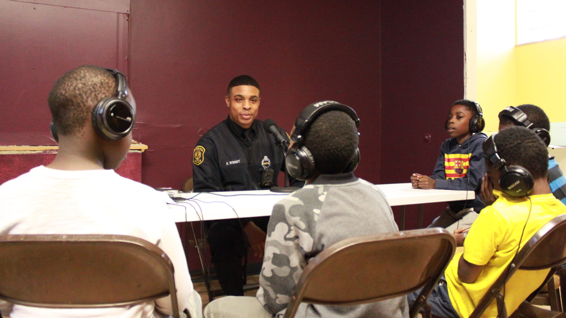 Five children wearing headphones interviewing an adult wearing a police uniform with a microphone connected to a portable recorder