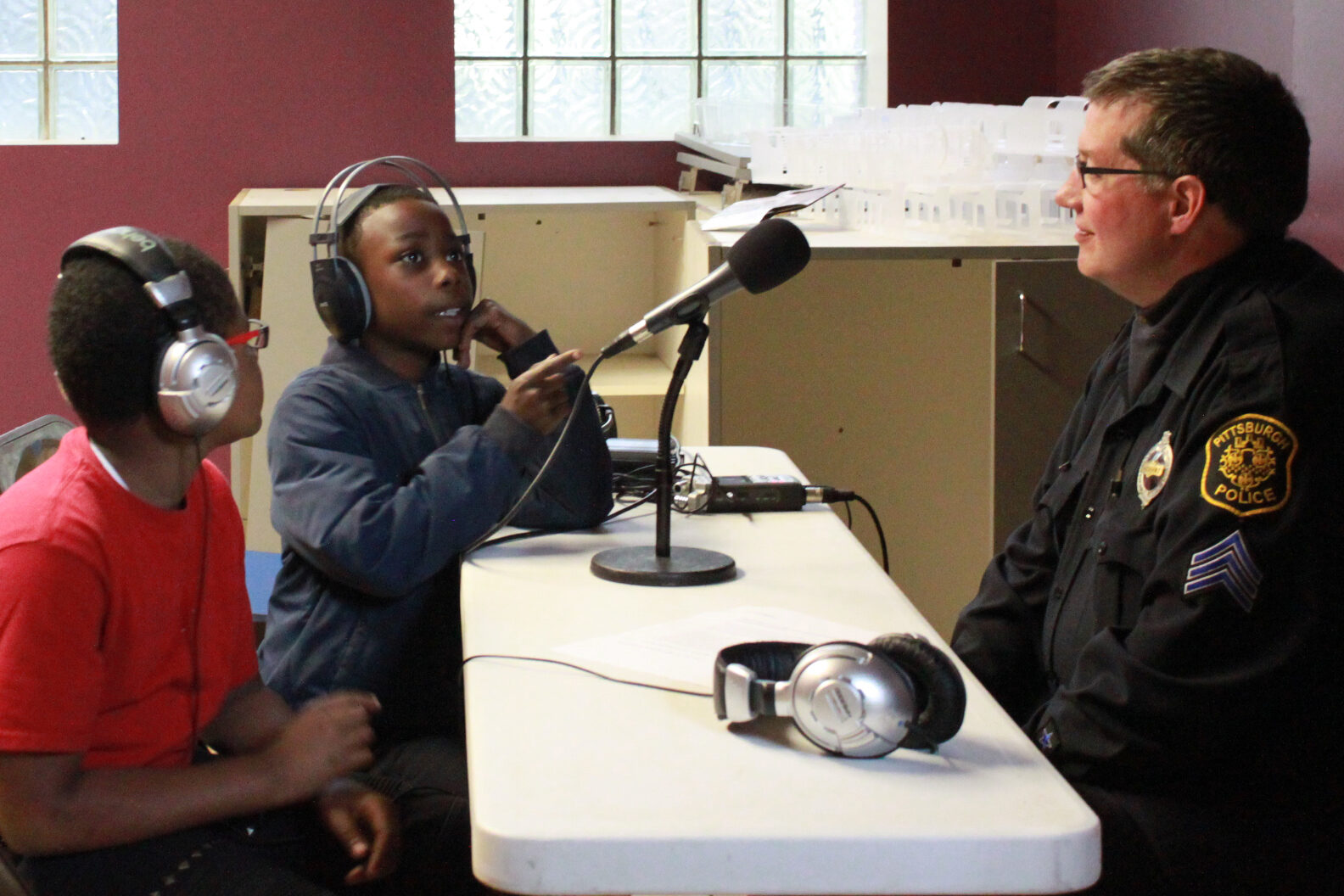 Two youth wearing headphones interviewing an adult wearing a police uniform with a microphone connected to a portable recorder