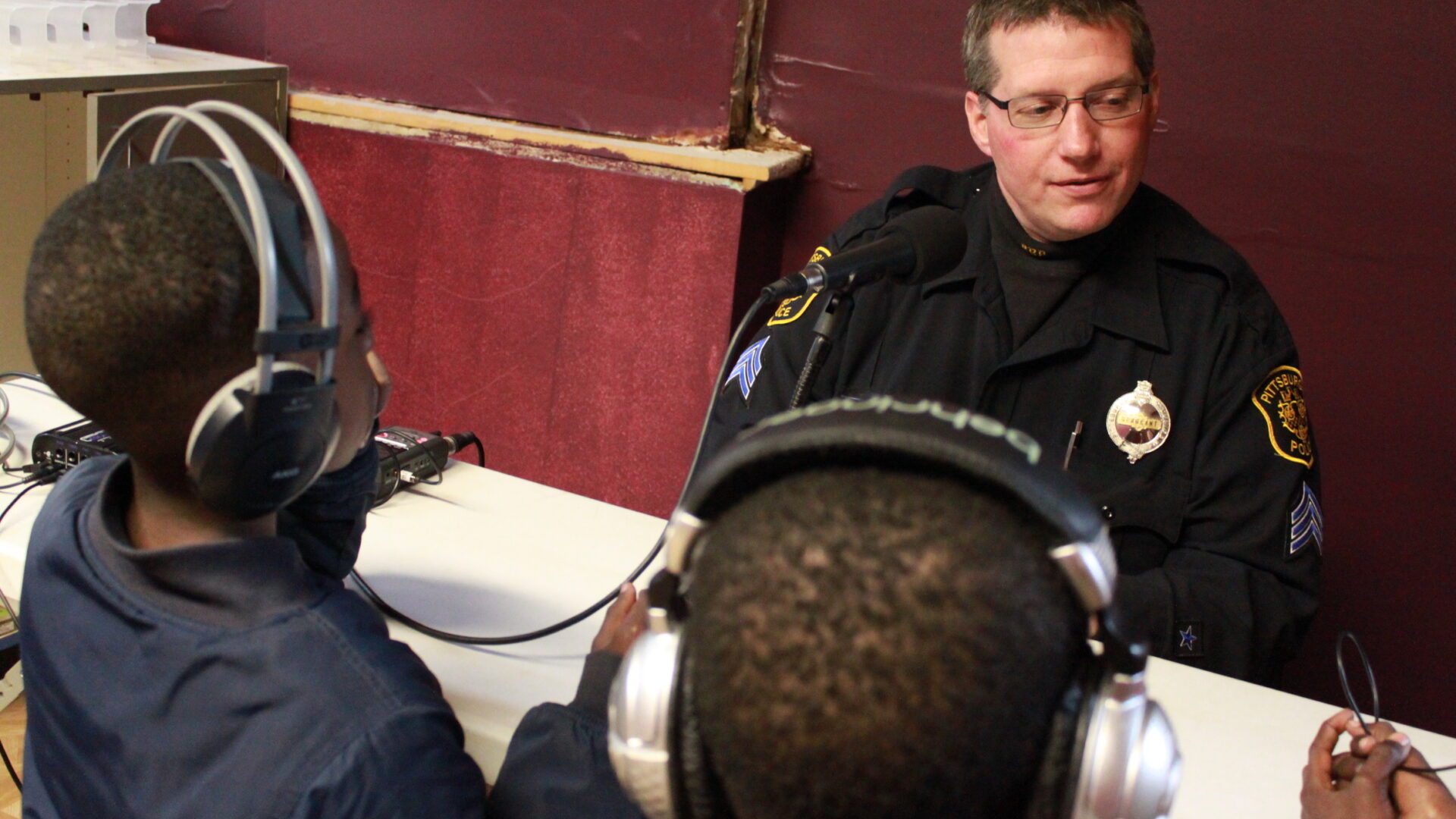 Two youth wearing headphones interviewing an adult with a microphone attached to a portable recorder