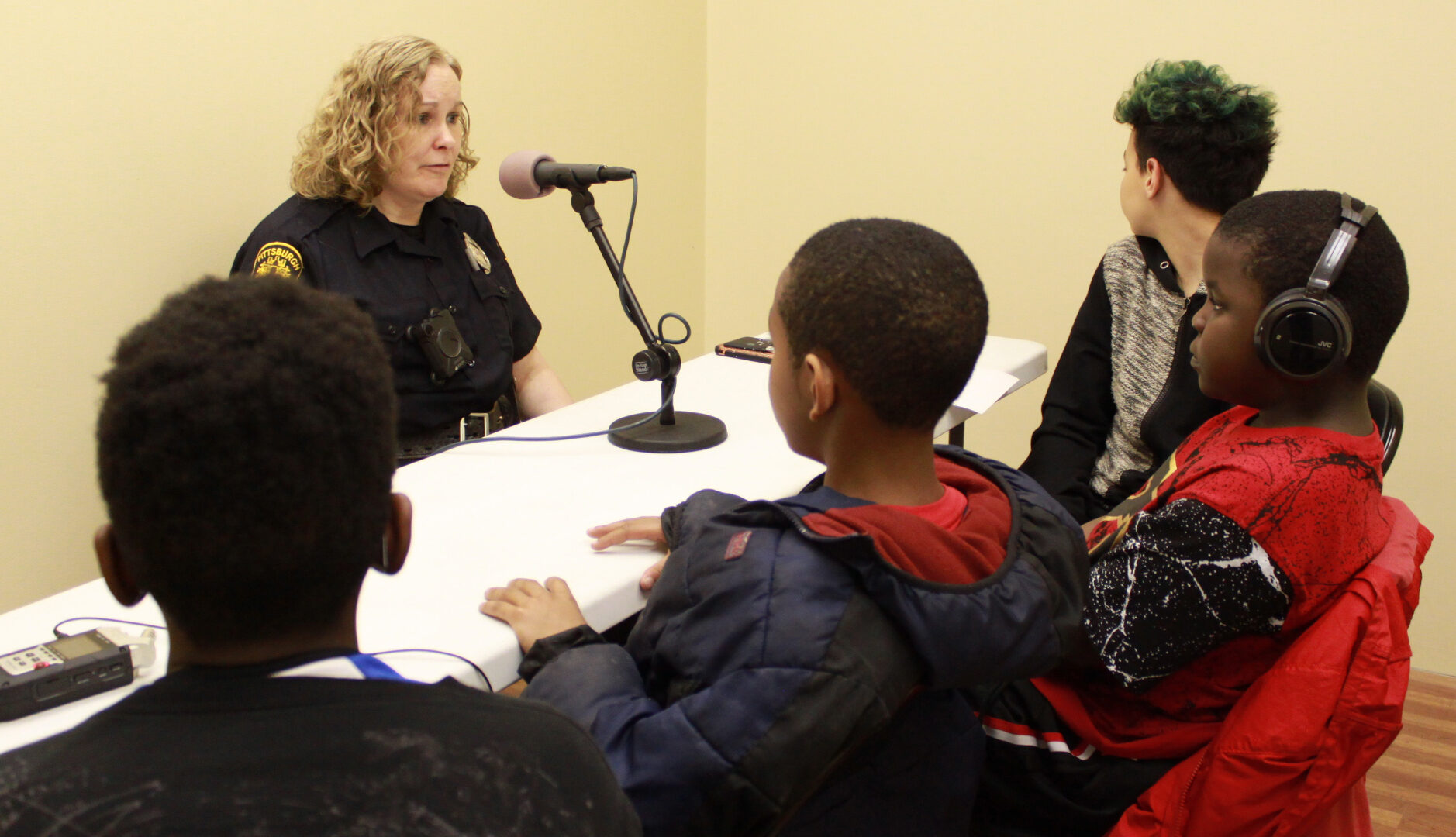 Four youth, one wearing headphones, interviewing an adult wearing a police uniform with a microphone attached to a portable recorder