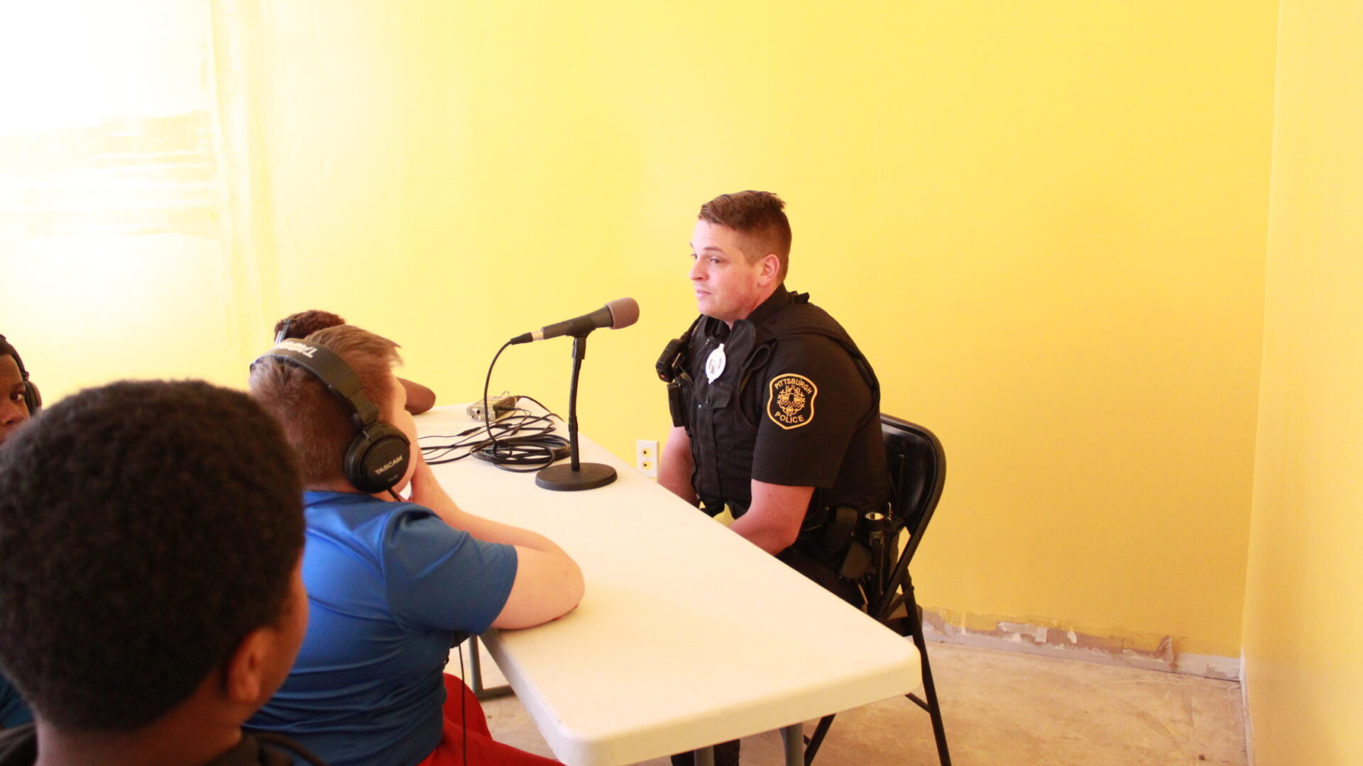 Four children, three wearing headphones, using a portable recorder and a microphone to interview an adult wearing a police uniform