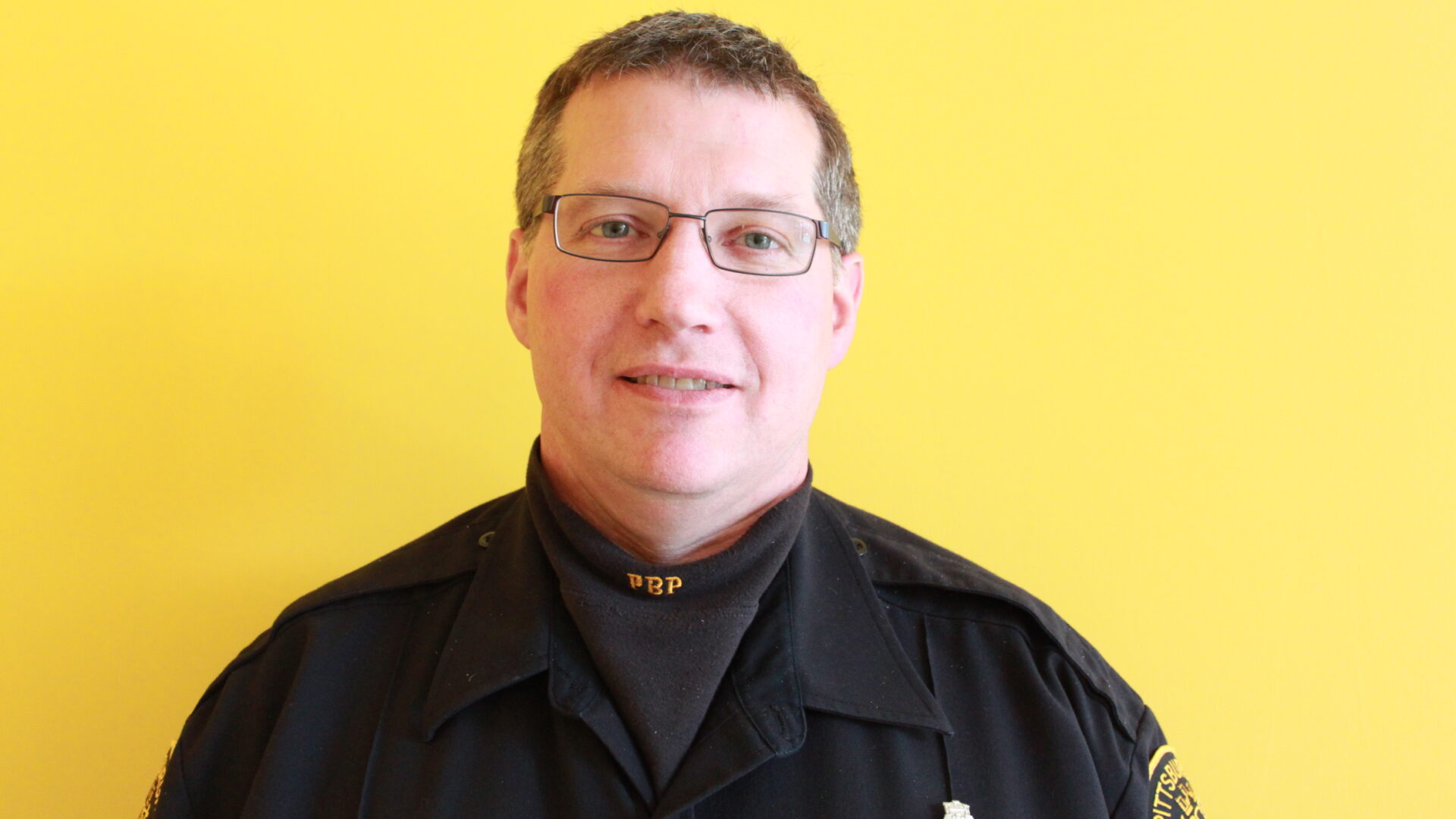Adult with short light brown hair wearing glasses and a police uniform smiling and posing in profile
