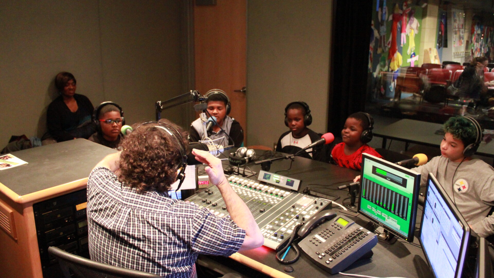 An adult wearing headphones and sitting behind a microphone and a mixing board interviewing five youth wearing headphones sitting behind microphones in a radio studio