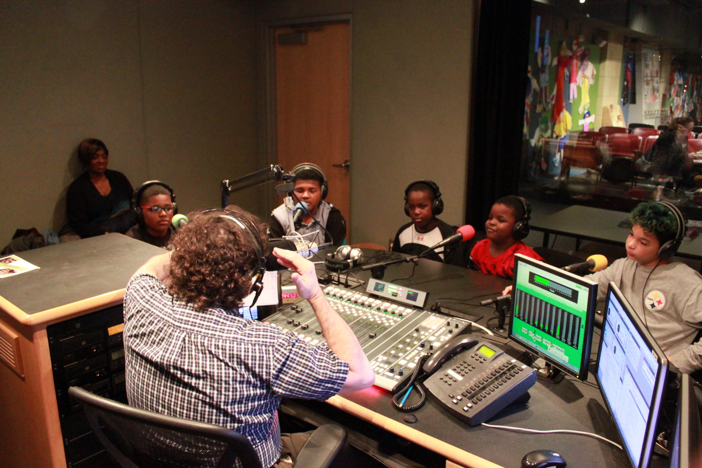 An adult wearing headphones and sitting behind a microphone and a mixing board interviewing five youth wearing headphones sitting behind microphones in a radio studio