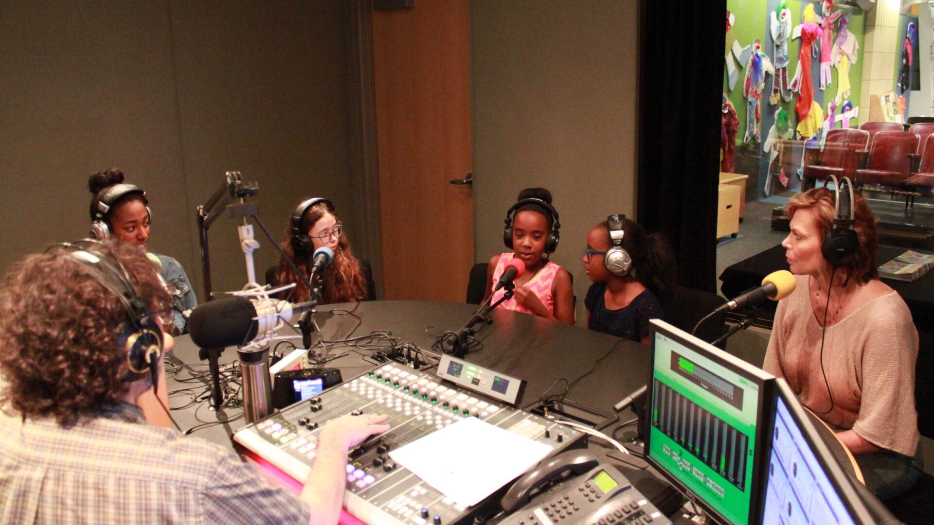 Four youth and an adult wearing headphones speaking into microphones being interviewed by an adult wearing headphones sitting behind a microphone and a mixing board in a radio studio