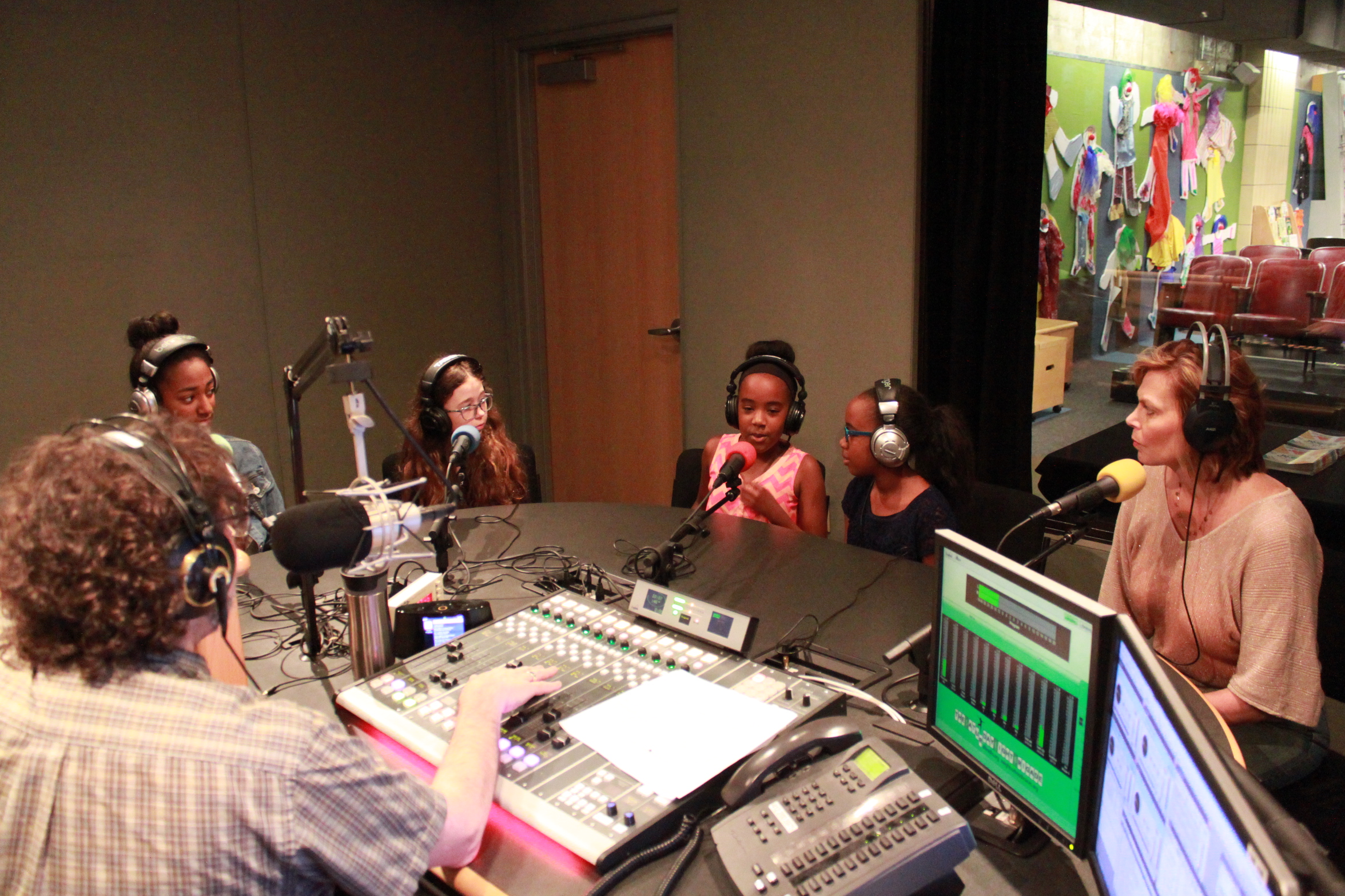 Four youth and an adult wearing headphones speaking into microphones being interviewed by an adult wearing headphones sitting behind a microphone and a mixing board in a radio studio