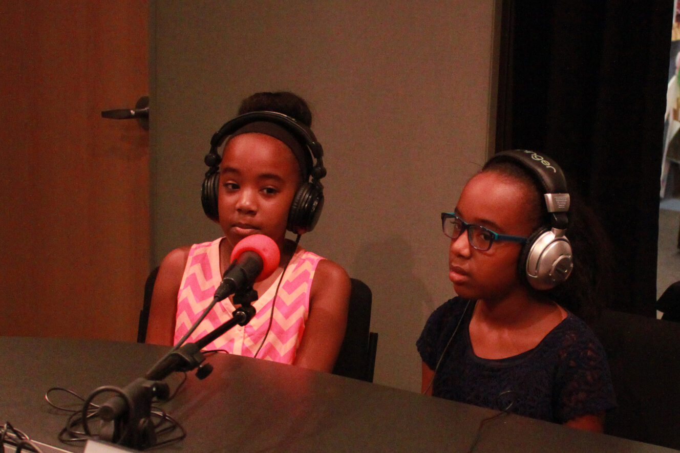 Two youth wearing headphones sitting in front of a microphone in a radio studio