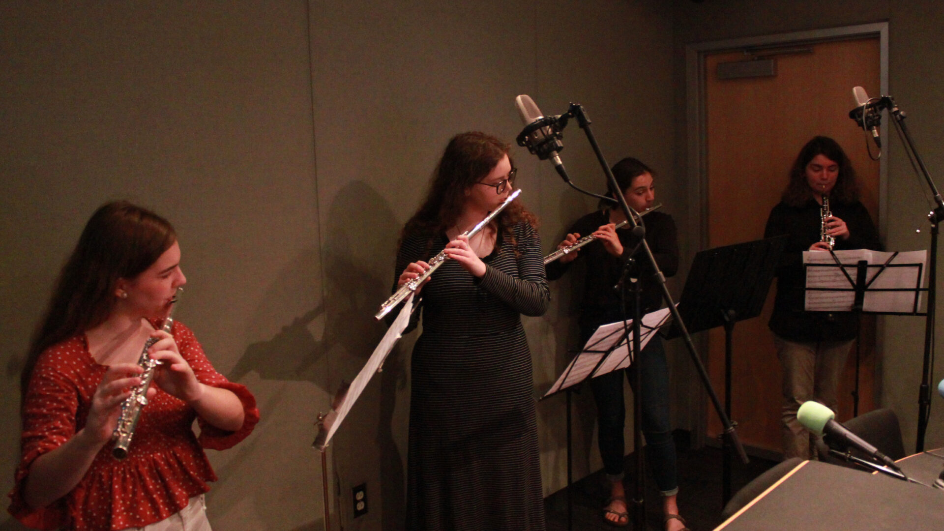 Three teens playing flute and one teen playing oboe, all looking at sheet music on music stands in a radio studio