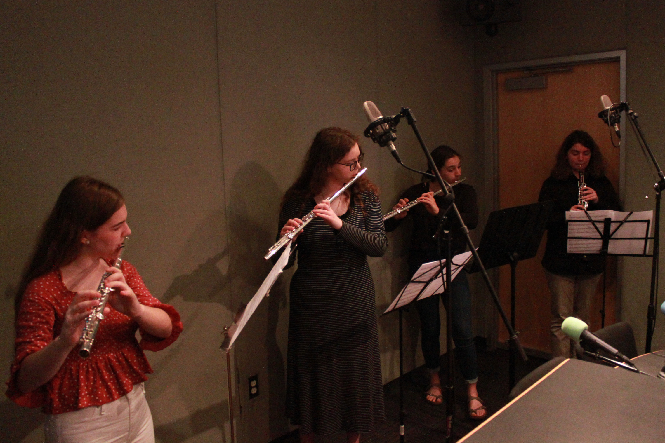 Three teens playing flute and one teen playing oboe, all looking at sheet music on music stands in a radio studio