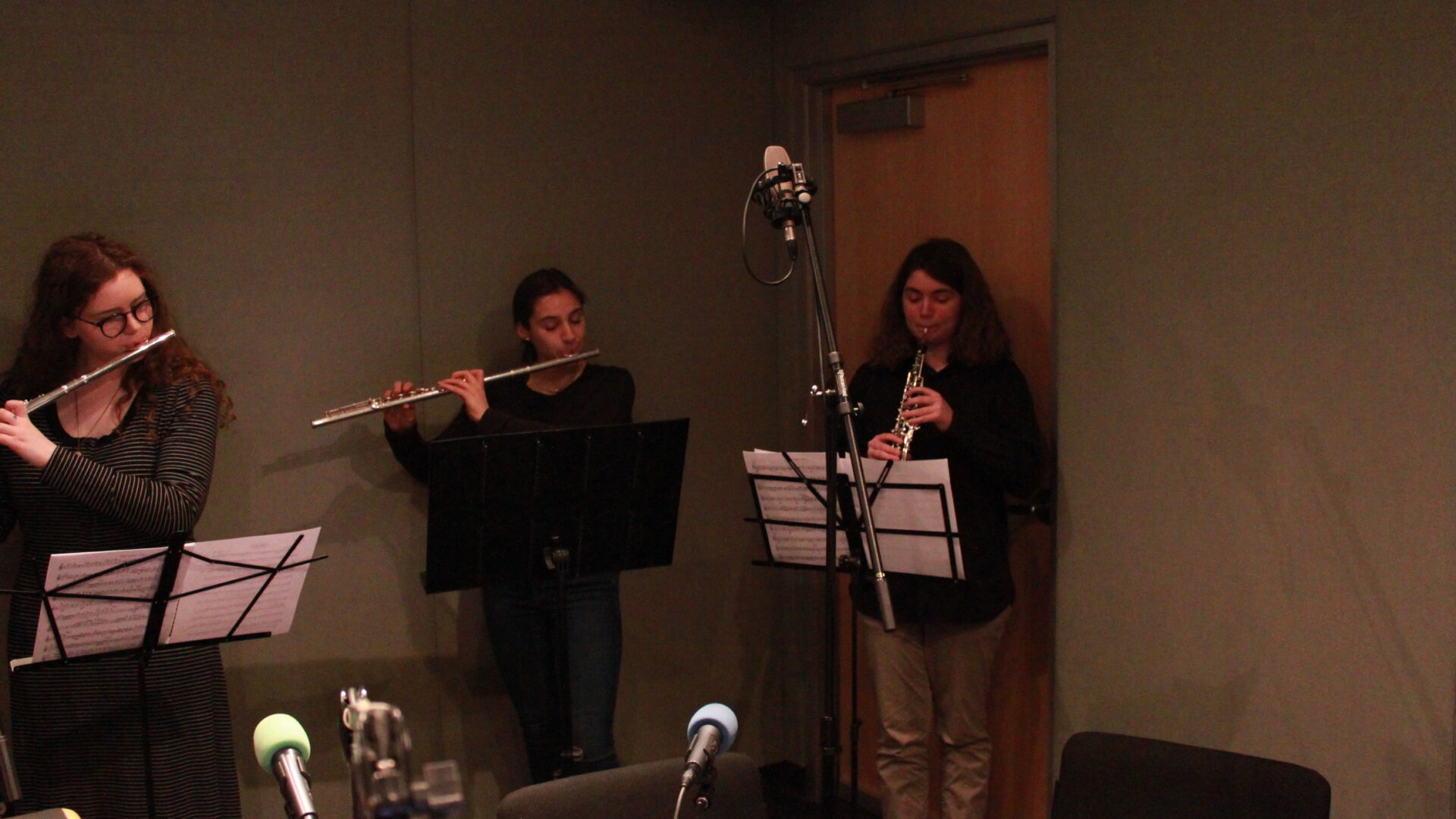Three teens performing on flutes and oboe looking at sheet music on music stands in a radio studio