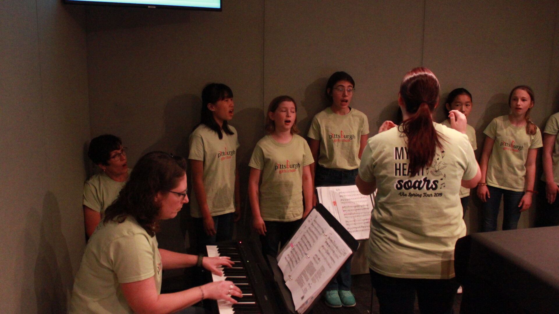 An adult playing a piano keyboard and another adult conducting five youth and an adult singing in a radio studio