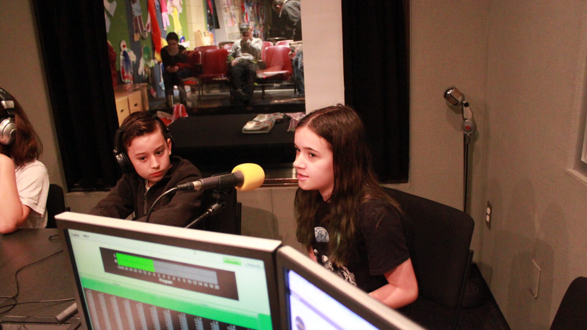 Two youth, one wearing headphones, speaking into a microphone in a radio studio