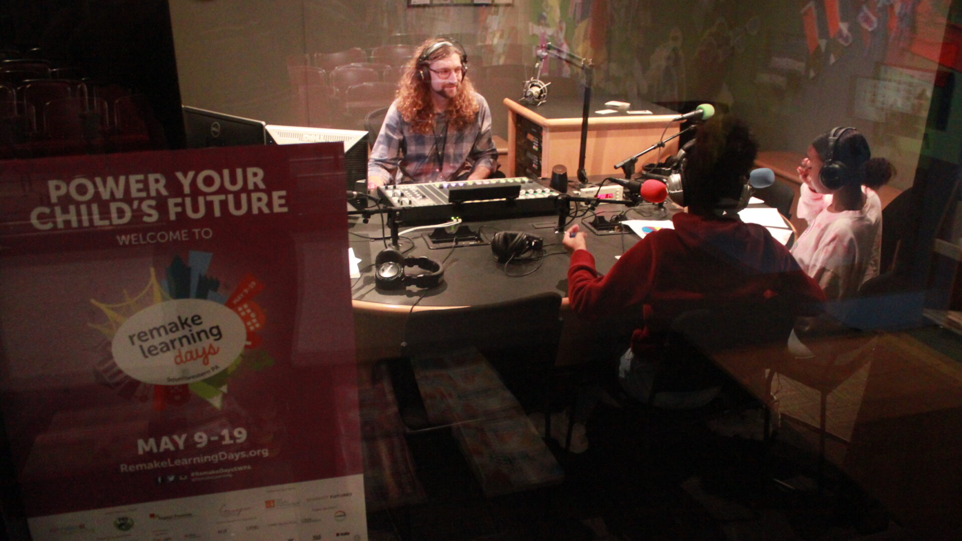 Two youth wearing headphones wearing headphones sitting in front of microphones looking at an adult wearing headphones sitting behind a mixing board in a radio studio
