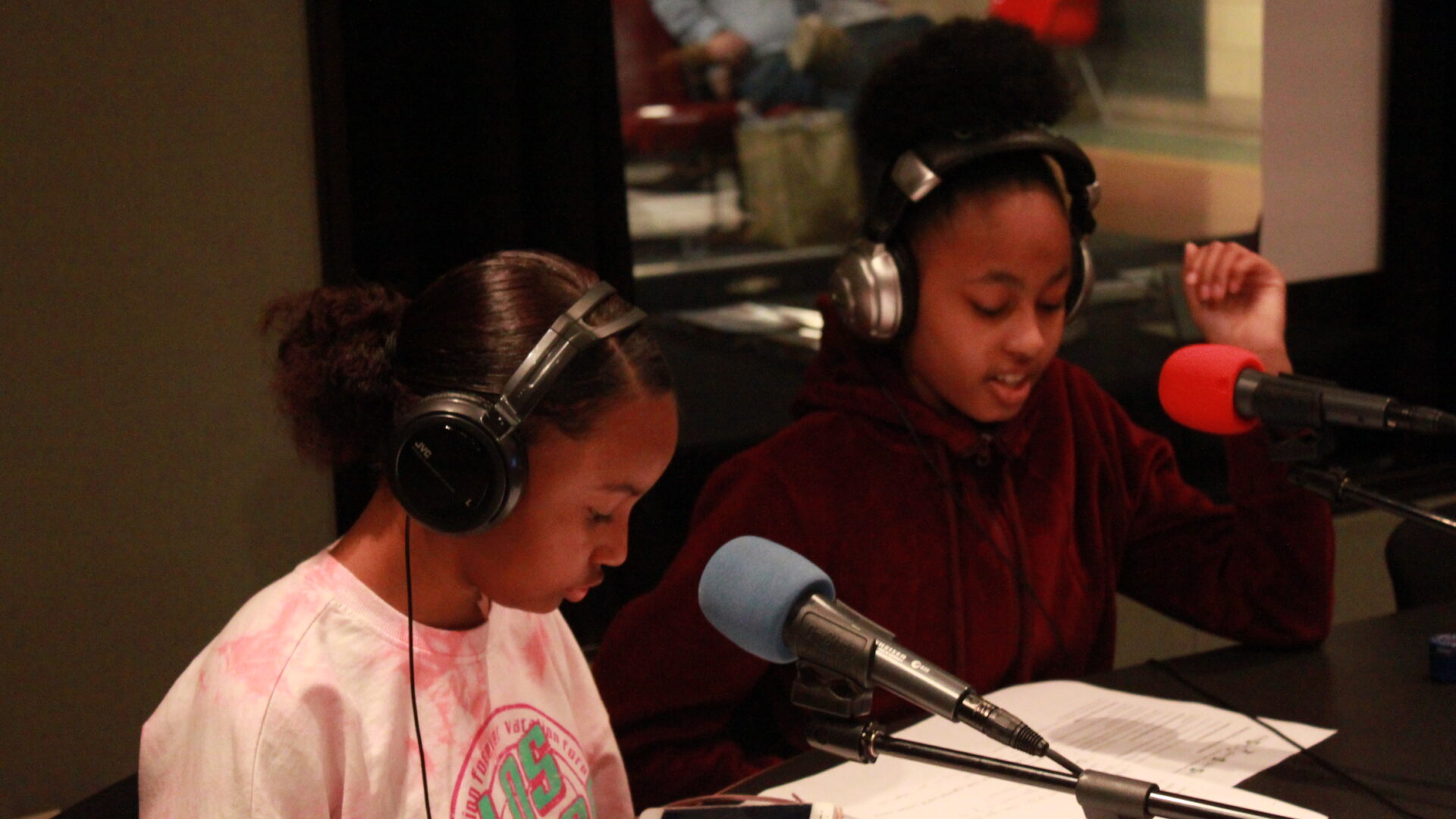Two teens wearing headphones and speaking into microphones looking down at pieces of paper and a smartphone in a radio studio