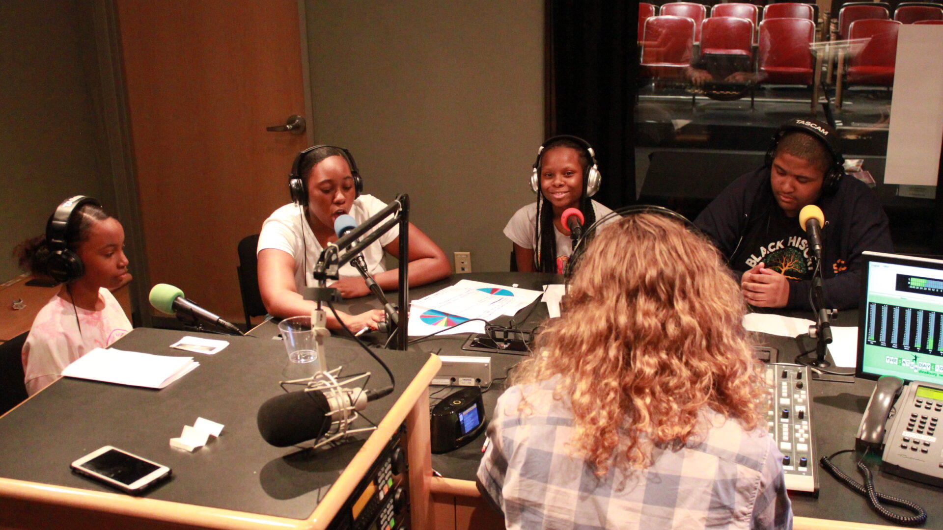 Four youth wearing headphones and speaking into microphones being interviewed by an adult wearing headphones in a radio studio