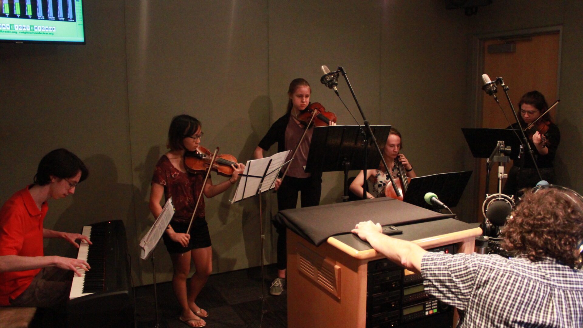 Five youth performing on piano keyboard, two violins, viola, and cello while looking at sheet music on music stands while an adult wearing headphones listens in a radio studio