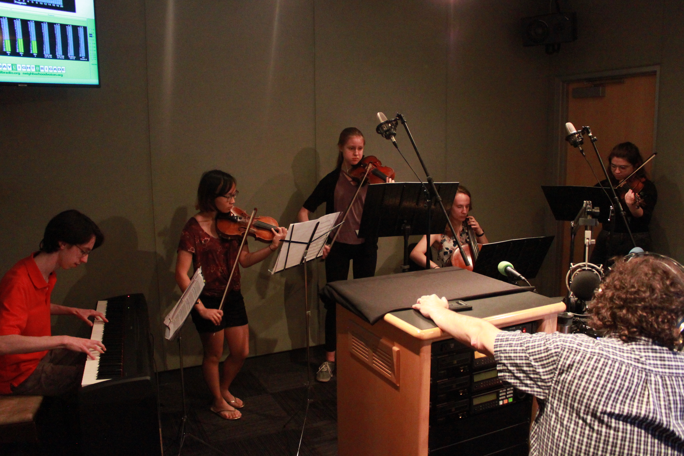 Five youth performing on piano keyboard, two violins, viola, and cello while looking at sheet music on music stands while an adult wearing headphones listens in a radio studio
