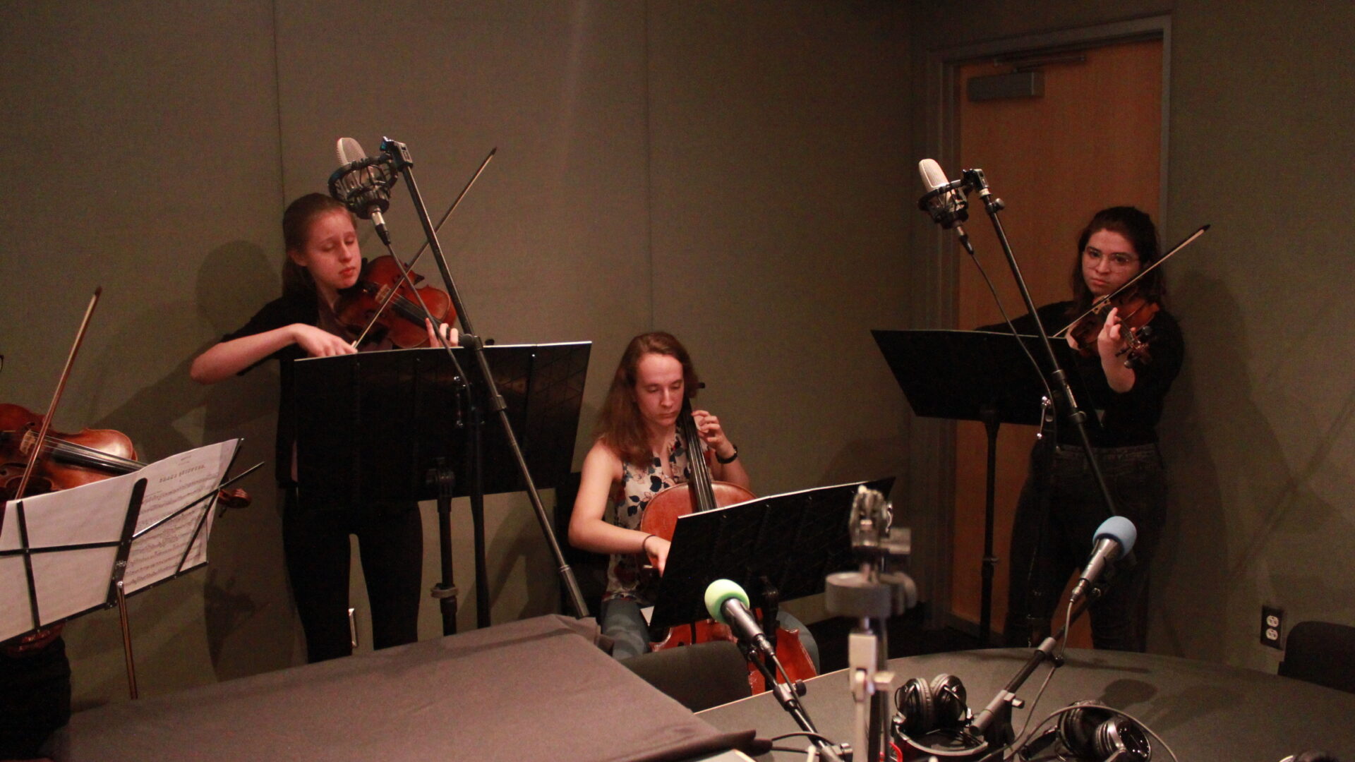 Four youth playing violins, viola, and cello while looking at sheet music on music stands in a radio studio