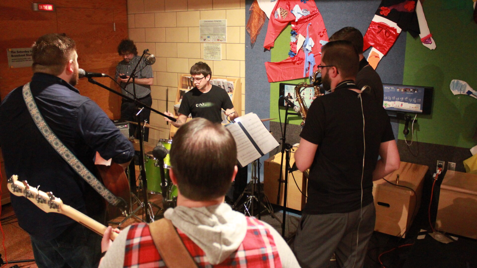 Five adults performing on alto saxophone, tenor saxophone, bass guitar, guitar, and drum set, the guitarist also singing into a microphone, with another adult in the background looking at their smartphone
