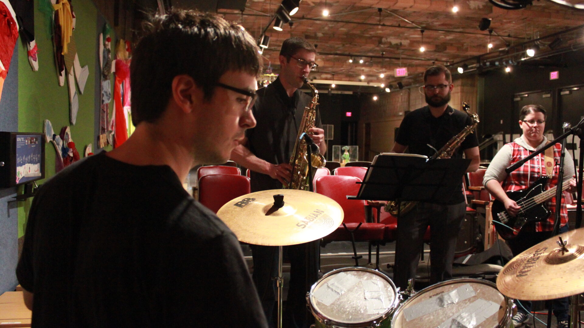 Four adults performing on drum set, alto saxophone, tenor saxophone, and bass