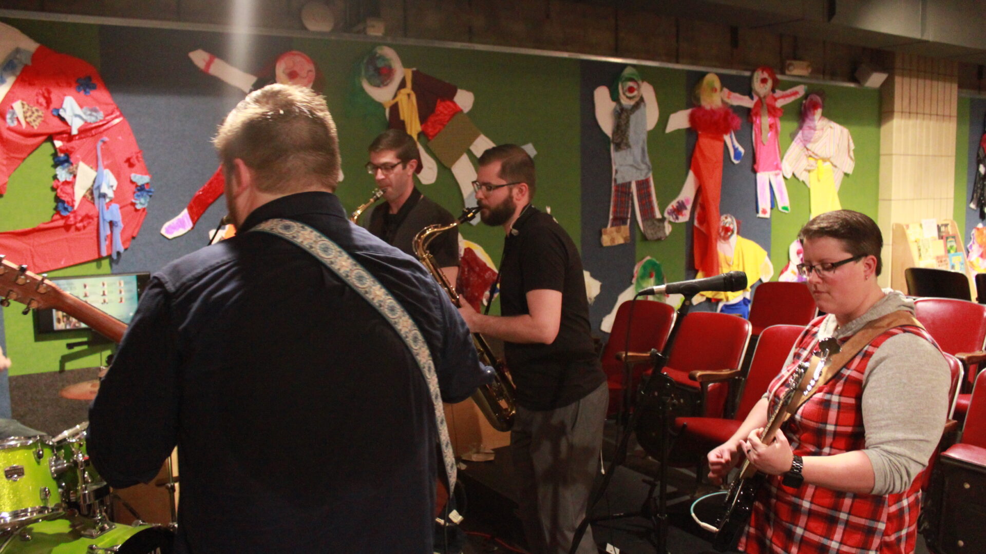 Four adults performing on alto saxophone, tenor saxophone, bass, and guitar, the guitarist also singing into a microphone