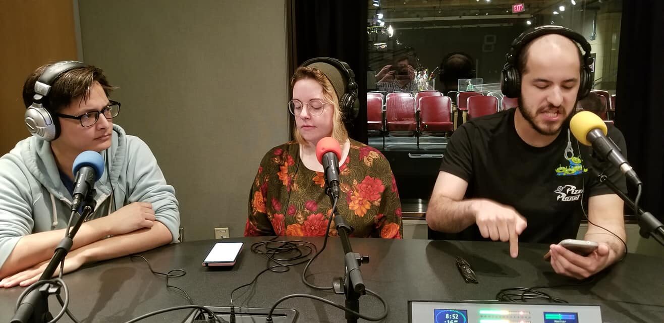 Three individuals wearing headphones and speaking into microphones and looking down at smartphones in a radio studio