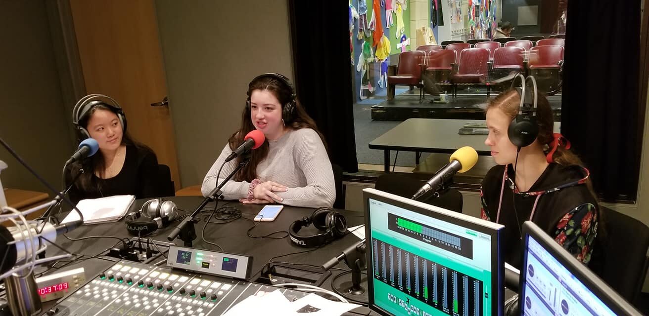 Three teens wearing headphones and speaking into microphones in a radio studio
