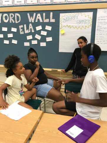 one youth speaking and holding a portable recorder while another youth wearing headphones listens while gesturing with their hands, and two other youth nearby observe in a classroom