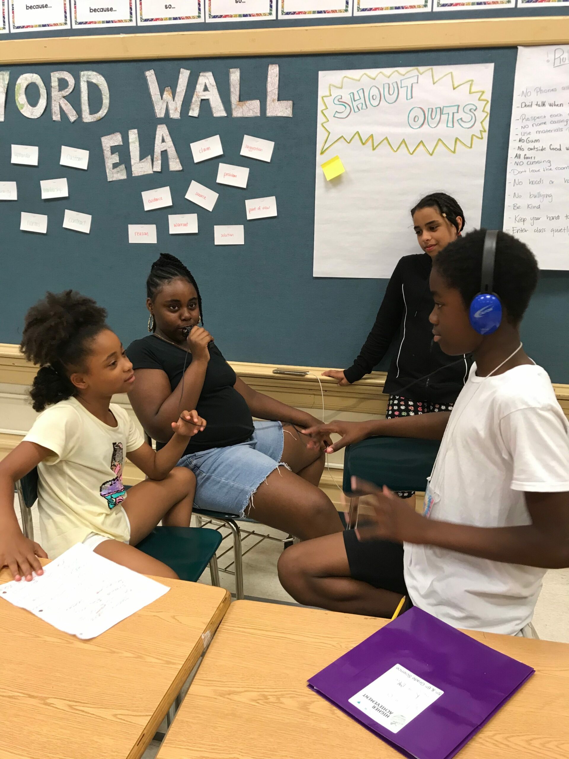 one youth speaking and holding a portable recorder while another youth wearing headphones listens while gesturing with their hands, and two other youth nearby observe in a classroom