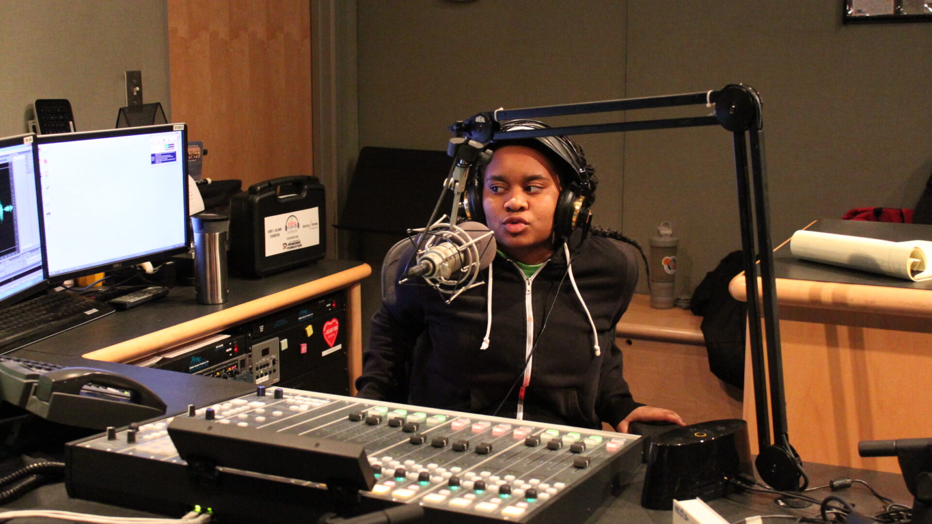 Teen wearing headphones sitting behind a microphone and a mixing board looking at a computer screen in a radio studio