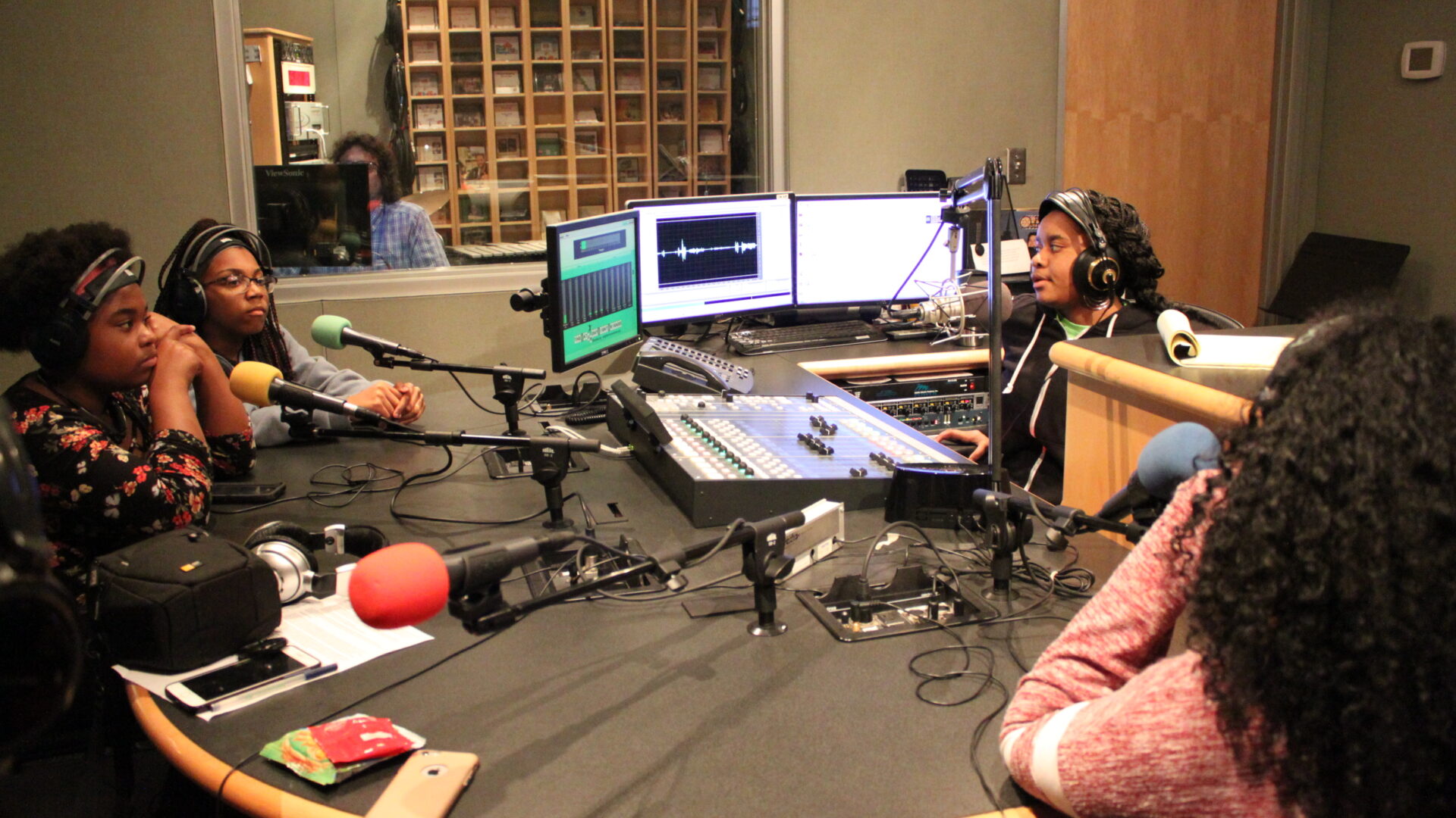 Four teens wearing headphones sitting behind microphones being interviewed by another teen wearing headphones sitting behind a microphone and a mixing board in a radio studio