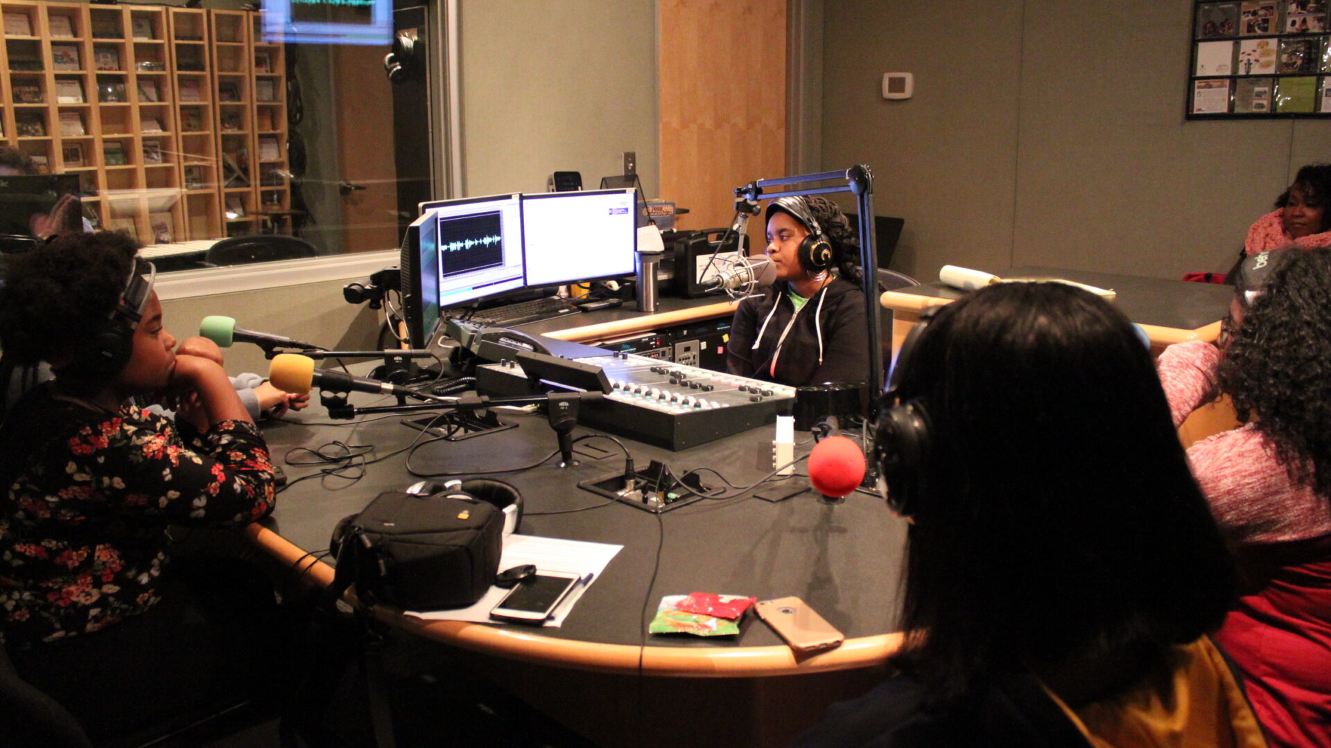 Four teens wearing headphones sitting behind microphones being interviewed by another teen wearing headphones sitting behind a mixing board and a microphone in a radio studio