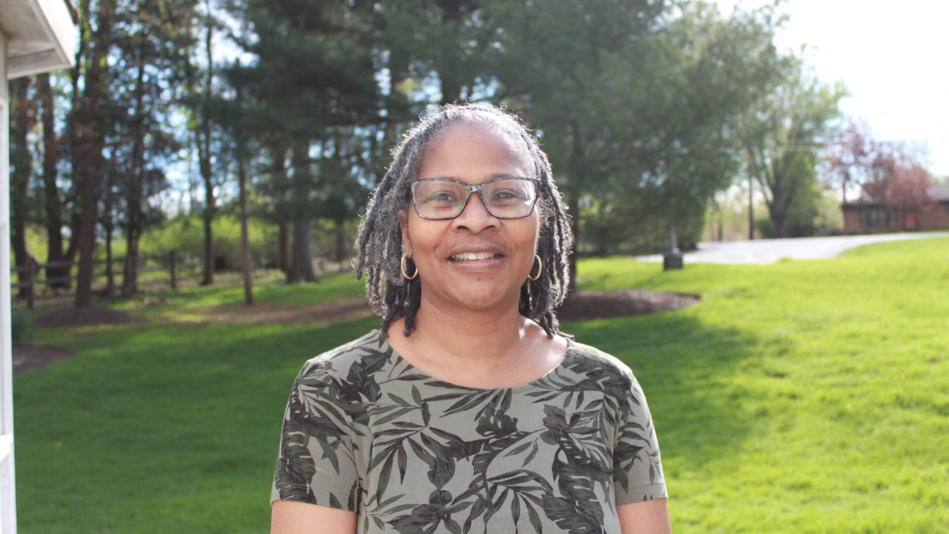 Adult wearing glasses smiling in profile in a large field of grass in front of a group of trees