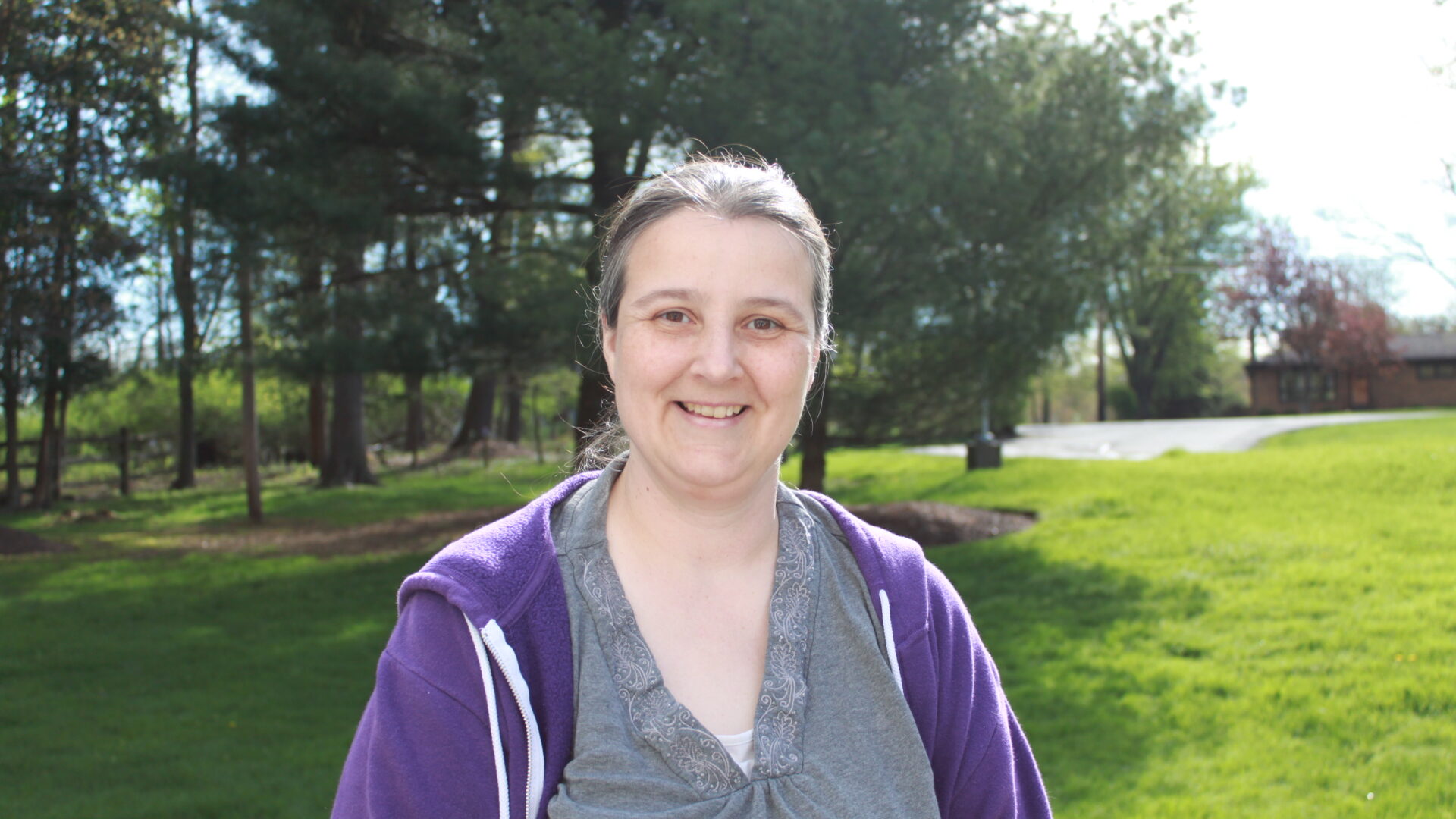 Adult smiling in profile in front of a field of grass and a group of trees