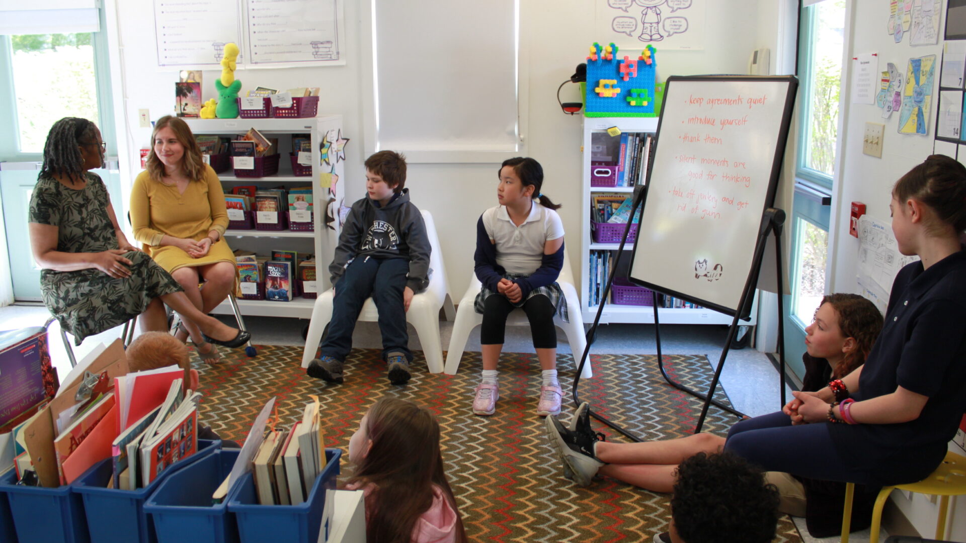 Six youth sitting around a multicolored striped rug and listening to two adults