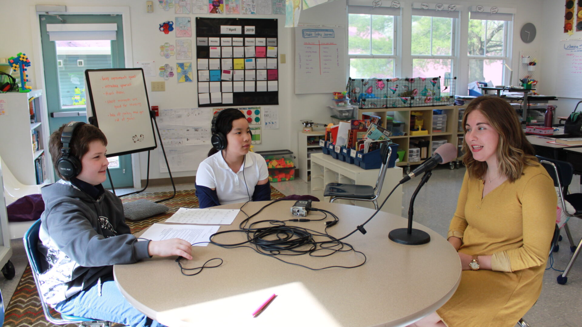 Two youth wearing headphones interviewing an adult speaking into a microphone connected to a portable recorder