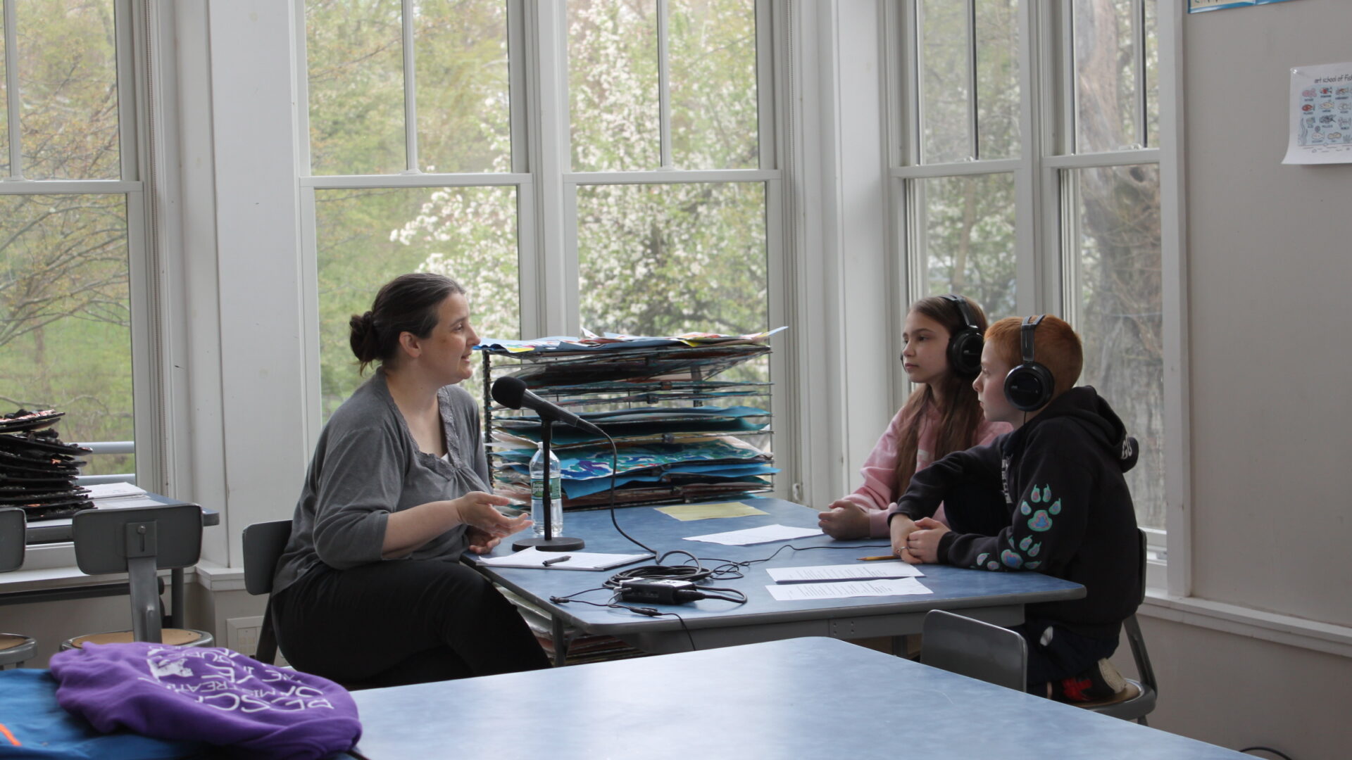 Two youth wearing headphones interviewing an adult speaking into a microphone connected to a portable recorder