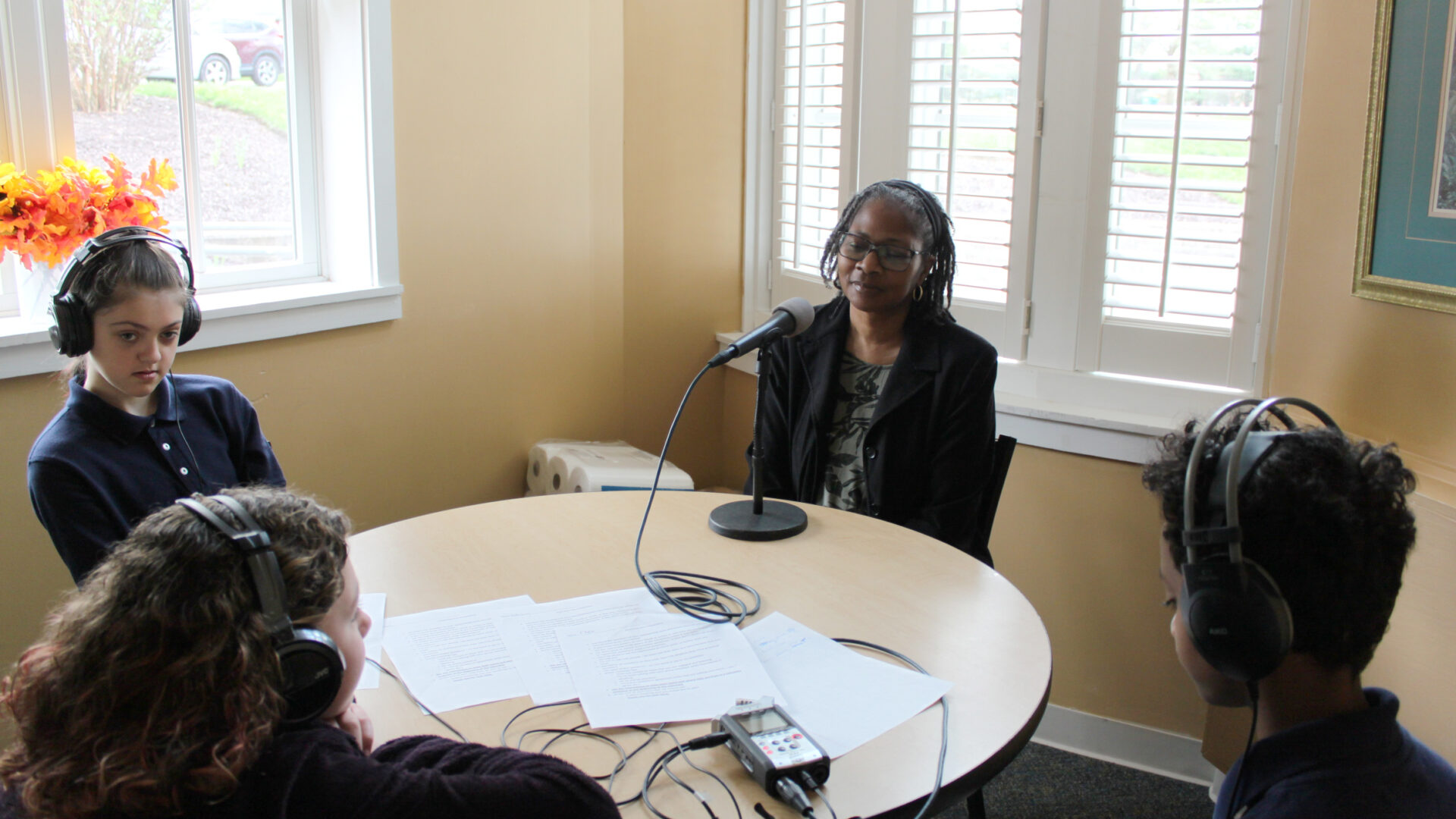 An adult sitting behind a microphone connected to a portable recorder being interviewed by three youth wearing headphones
