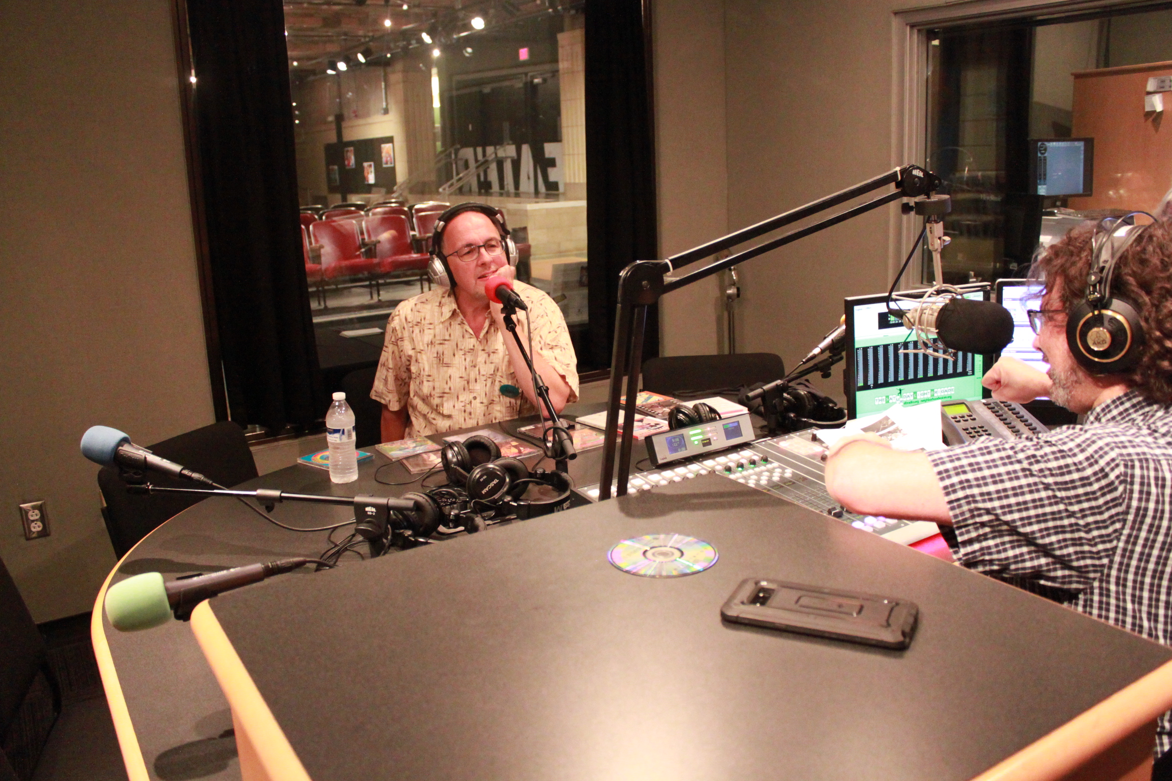 Adult wearing headphones and speaking into a microphone being interviewed by an adult wearing headphones and speaking into a microphone in a radio studio